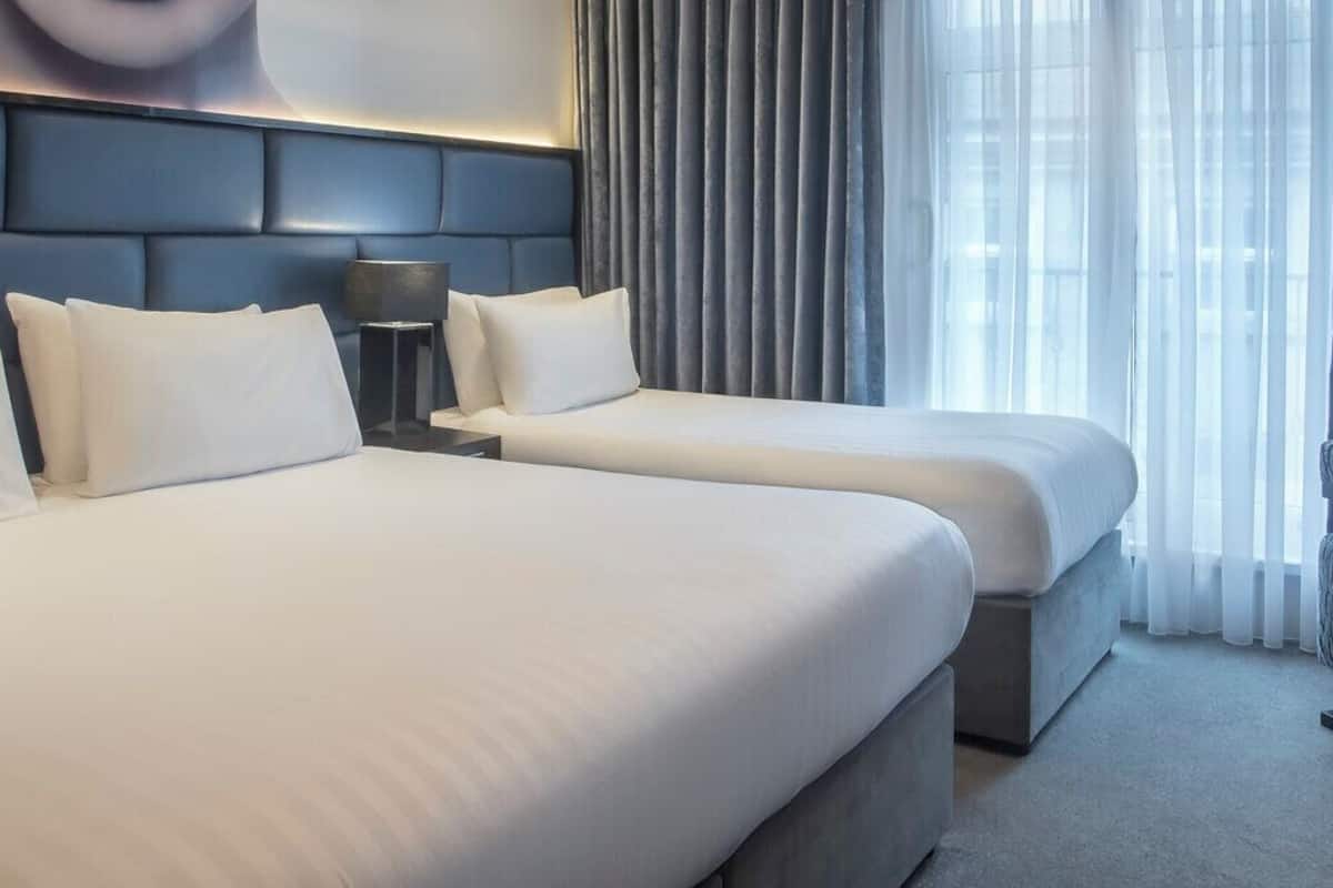 A hotel room with two beds, a headboard, a nightstand, a lamp, a chair, and a window with curtains.