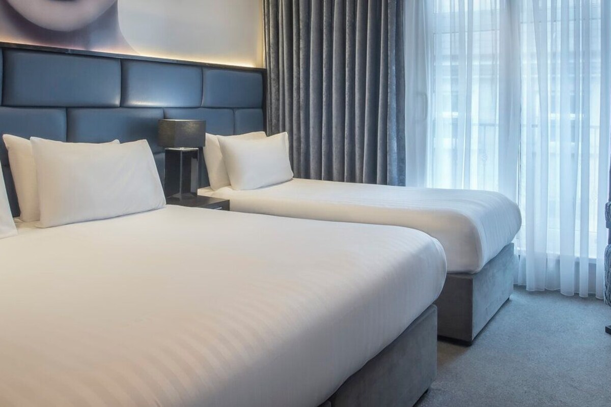 Una habitación de hotel con dos camas, cabecera, mesita de noche, lámpara, sillón y ventana con cortinas.