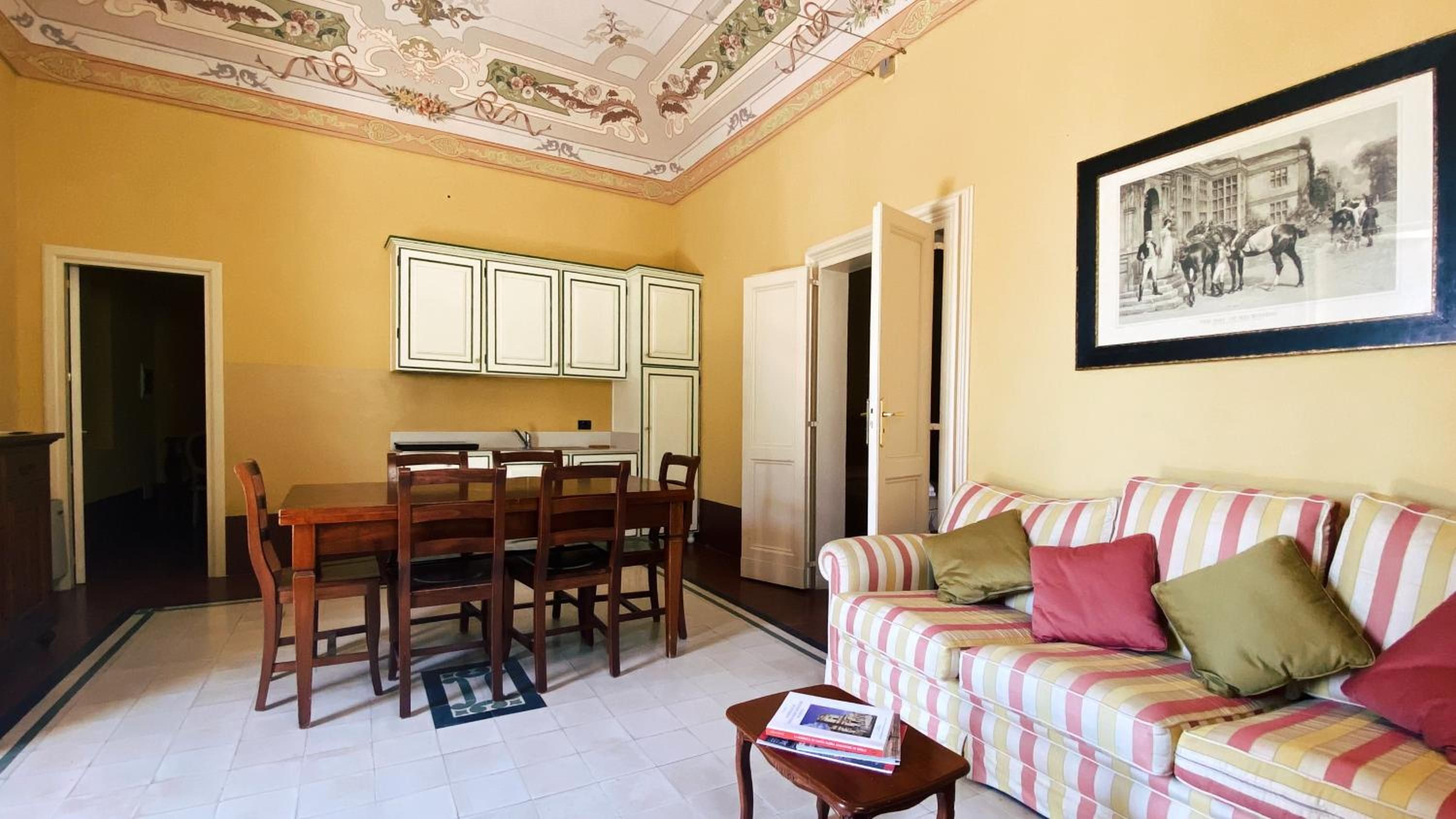 Una sala de estar con un sofá a rayas, una mesa de comedor con sillas, una zona de cocina con armarios y un cuadro en la pared.