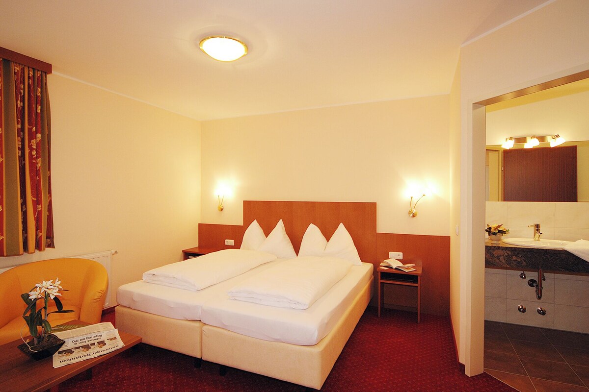 Una habitación de hotel con cama, mesita de noche, silla y baño con lavabo y espejo.