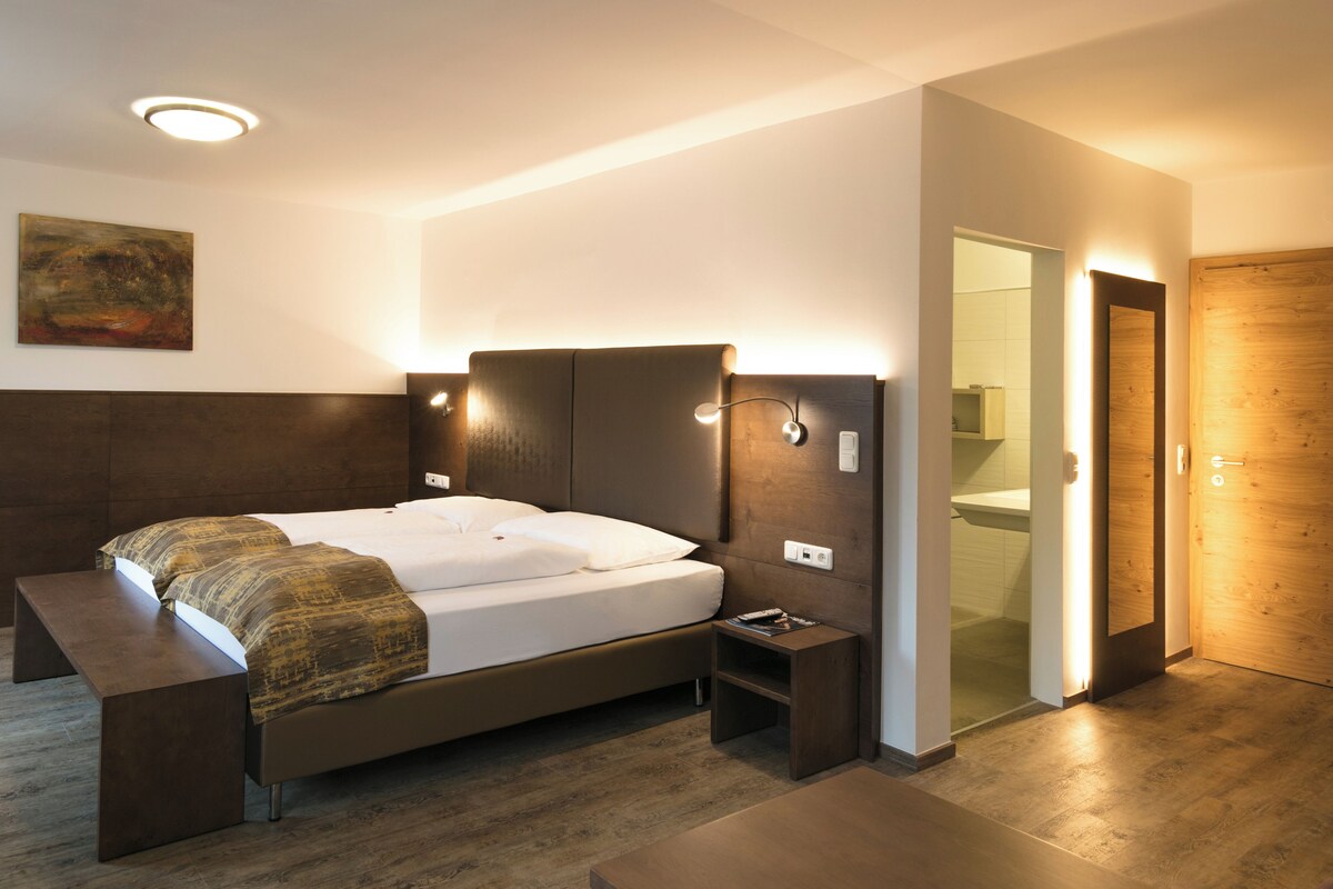 A modern hotel room with a large bed, bedside tables, a bench, a painting on the wall, and a bathroom visible through an open door.