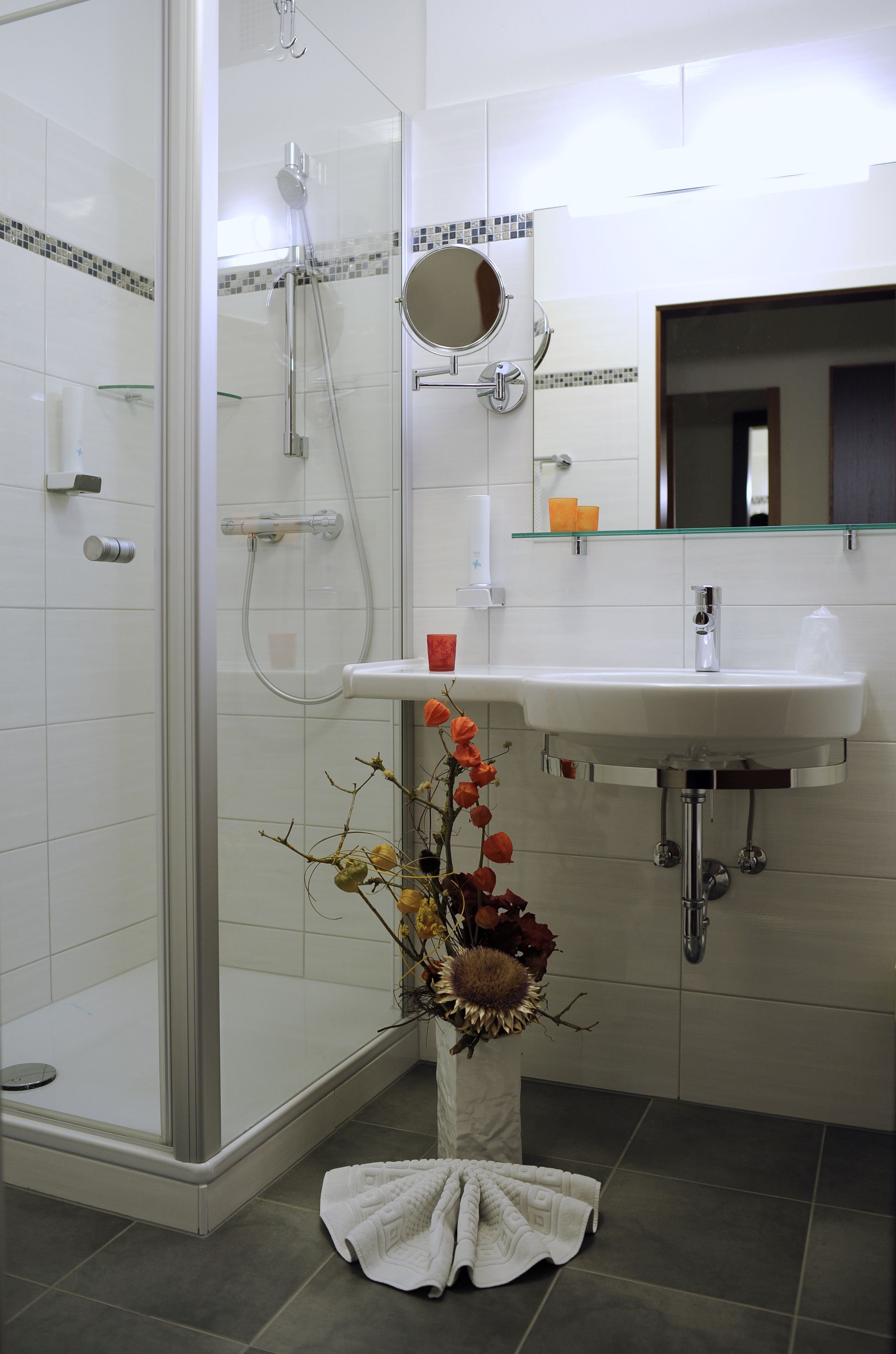 A modern bathroom with a glass shower enclosure, a white sink with a mirror above it, and a vase with dried flowers on the floor.