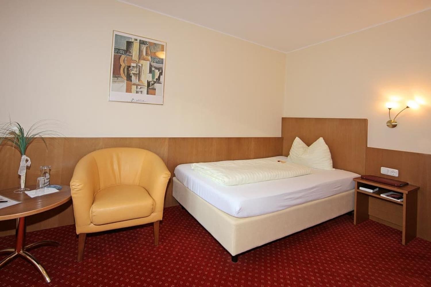 A hotel room with a bed, a chair, a small table, and a wall-mounted lamp.