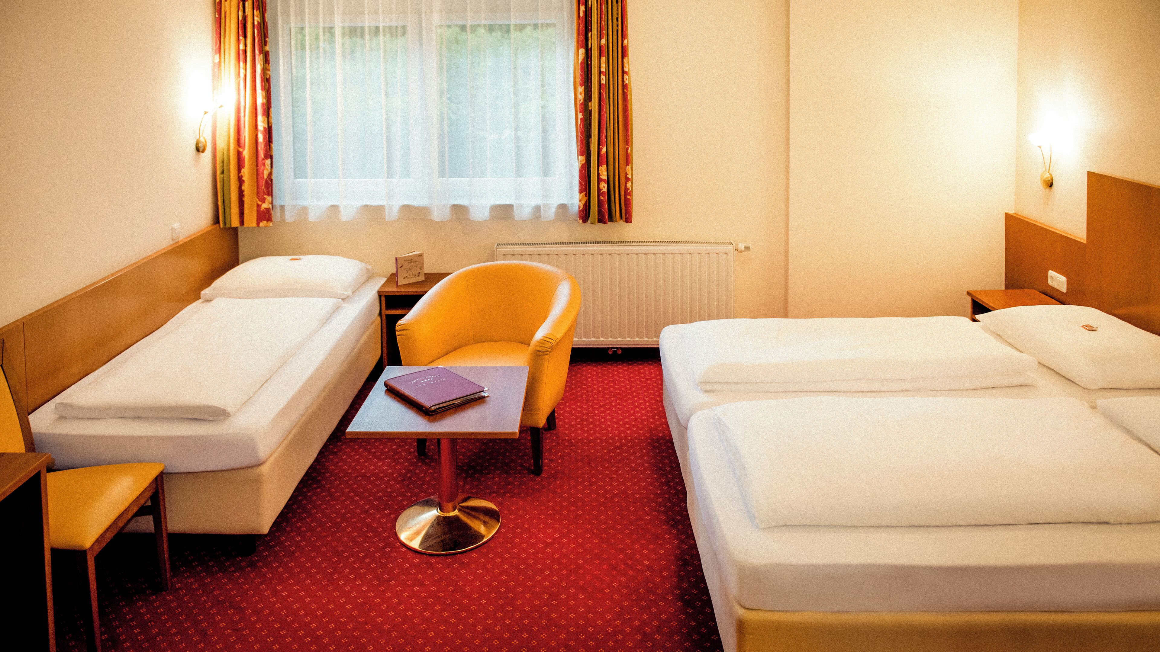 A hotel room with two single beds, a small table with a book, a chair, and a radiator.