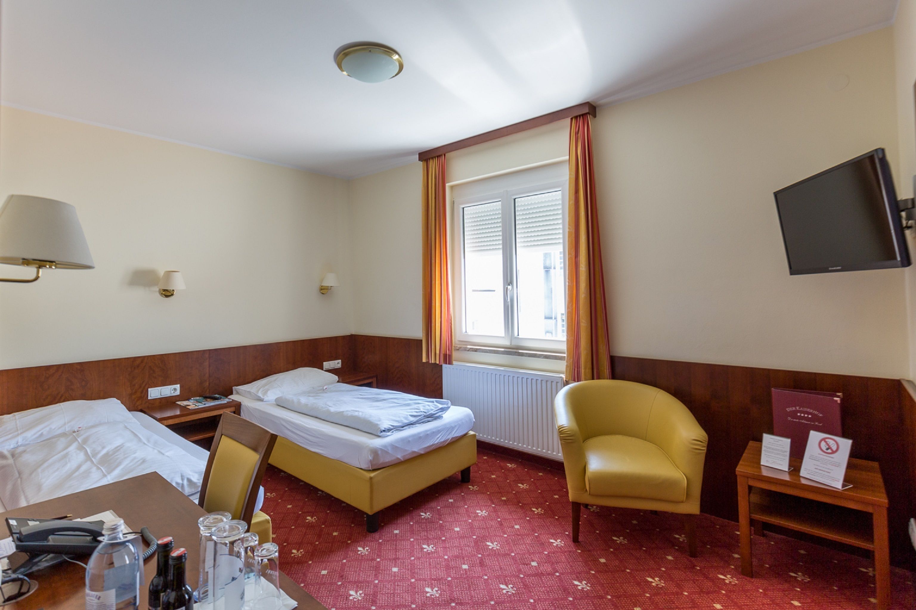 Ein Hotelzimmer mit einem Bett, einem Schreibtisch mit Telefon und Brille, einem Sessel, einem Fernseher und einem Fenster mit Vorhängen.