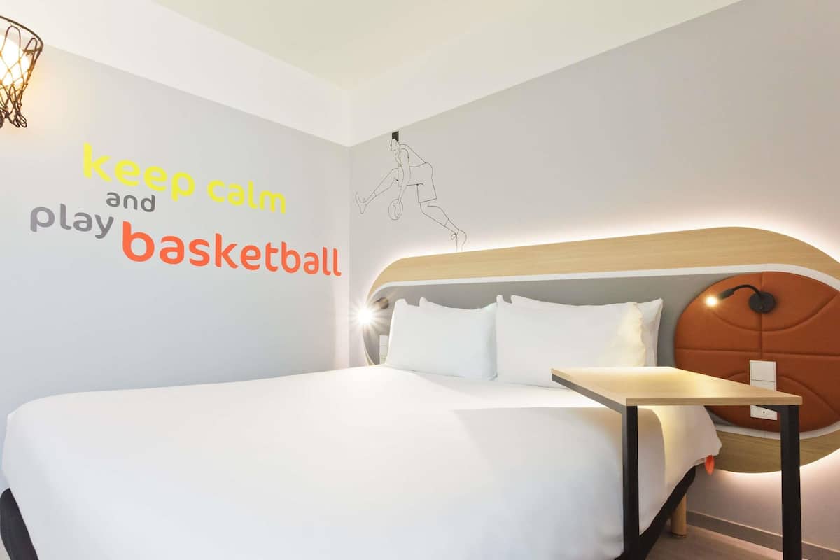 Una habitación de hotel moderna con una cama grande, una mesita de noche y una pared con texto motivacional e ilustración de un jugador de baloncesto.