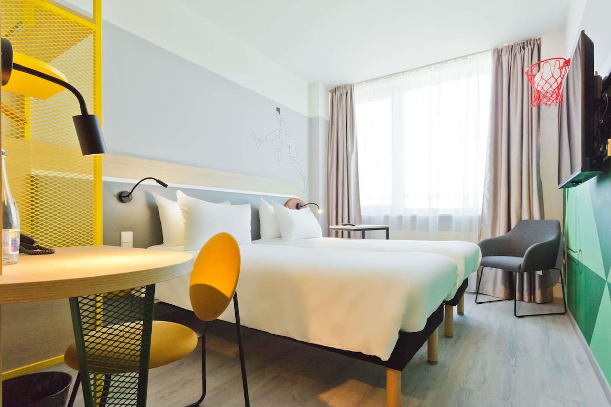 A hotel room with a bed, a desk with a chair, a TV, and a window with curtains.