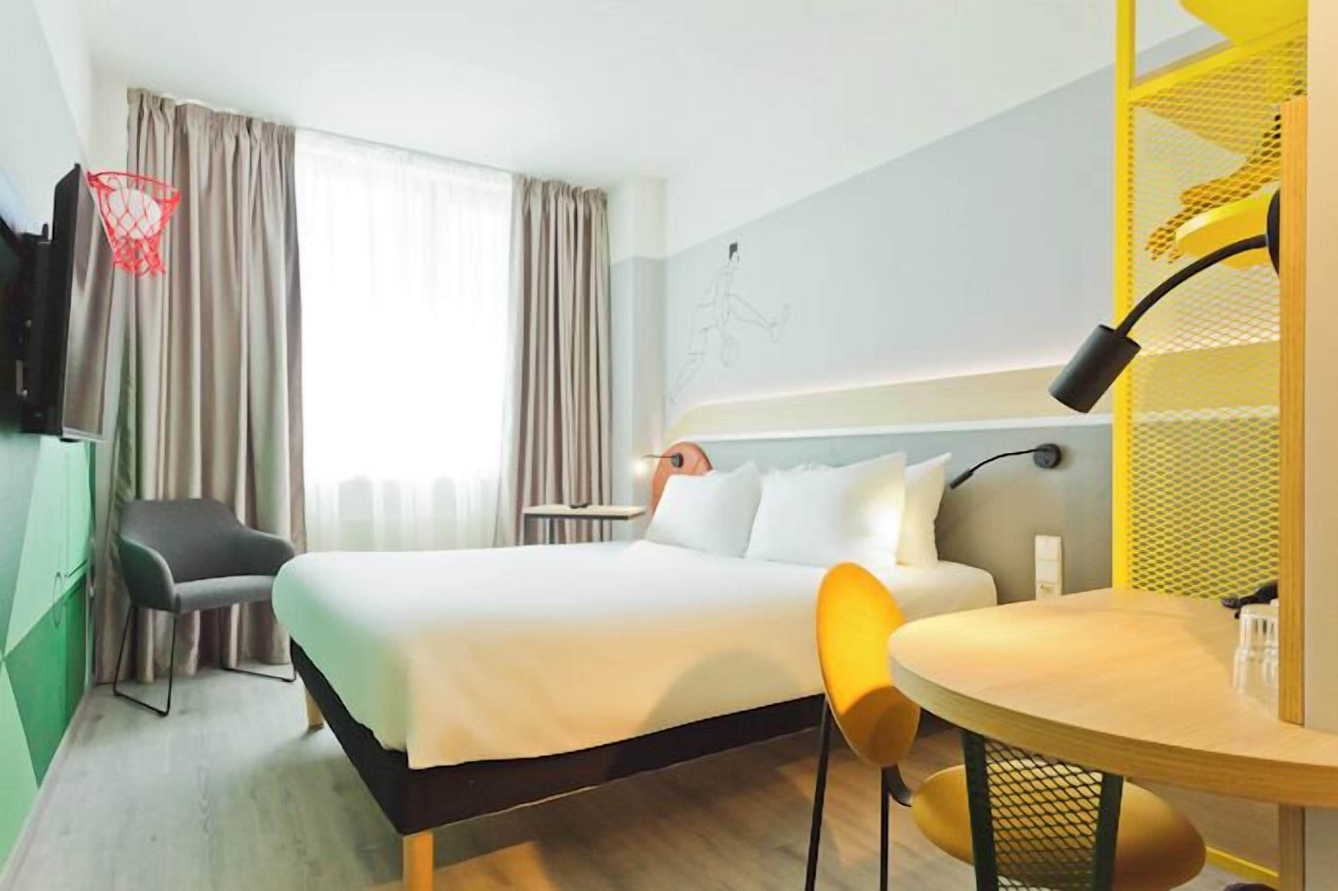 A hotel room with a bed, a desk, a chair, a TV, and a window with curtains.