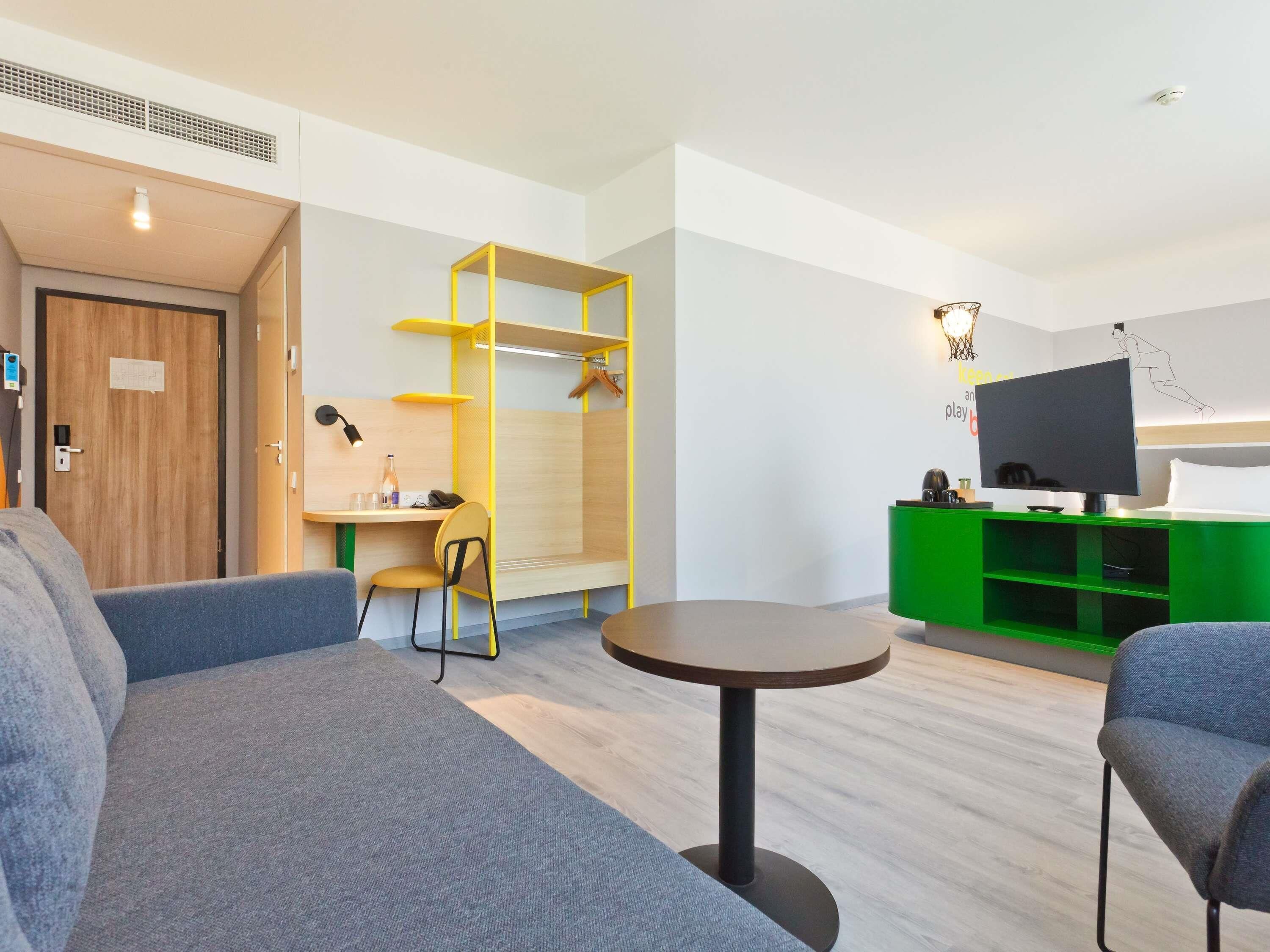 Une chambre d’hôtel moderne avec un canapé gris, une petite table ronde, un meuble de rangement vert et un téléviseur.