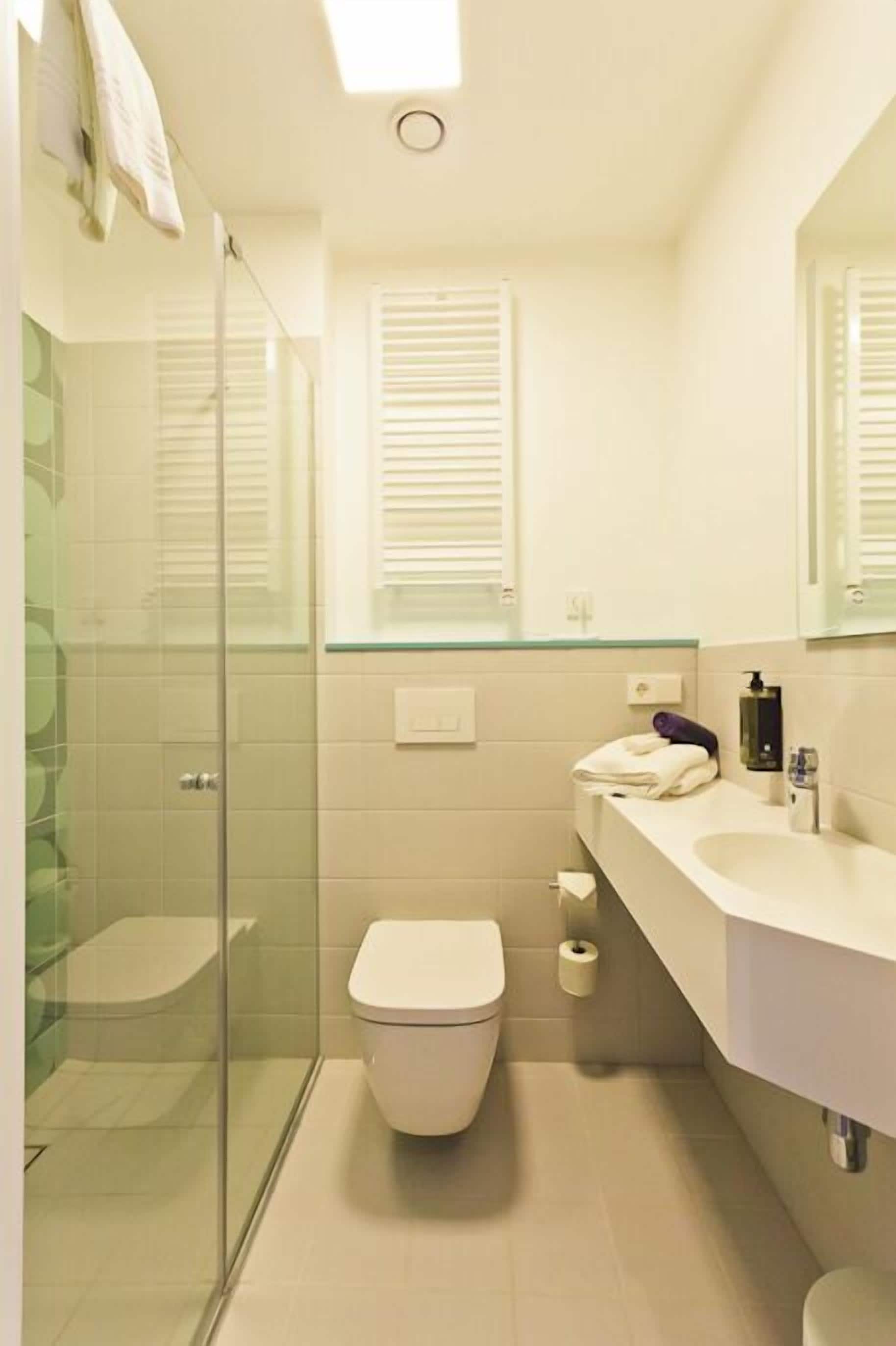 A bathroom with a glass shower enclosure, a toilet, a sink with a mirror, and a window with blinds.