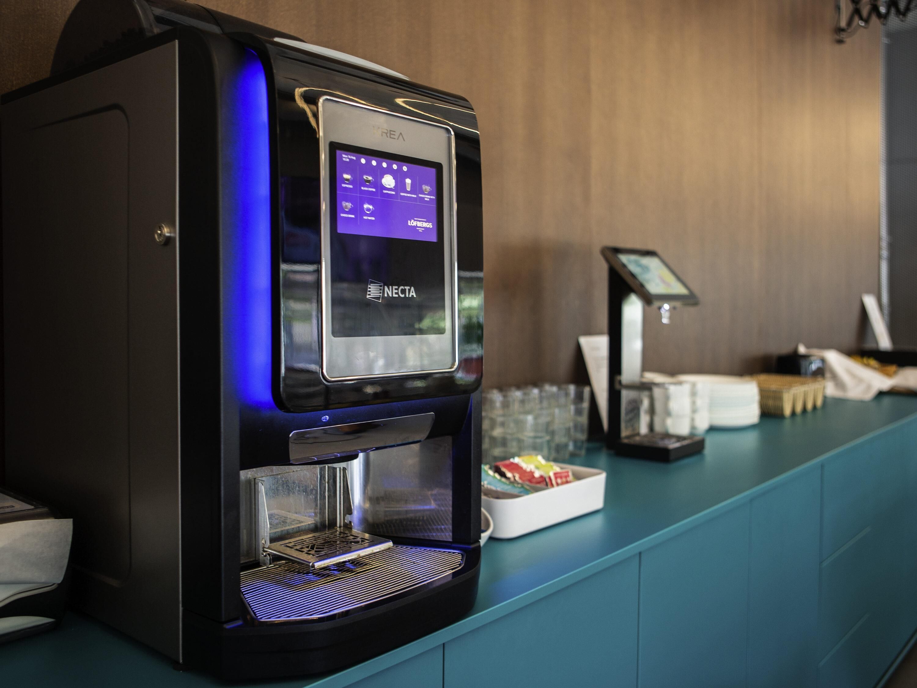 Une machine à café dotée d’une interface numérique, d’un distributeur d’eau et d’un présentoir rempli de gobelets jetables, le tout posé sur un comptoir de couleur turquoise.