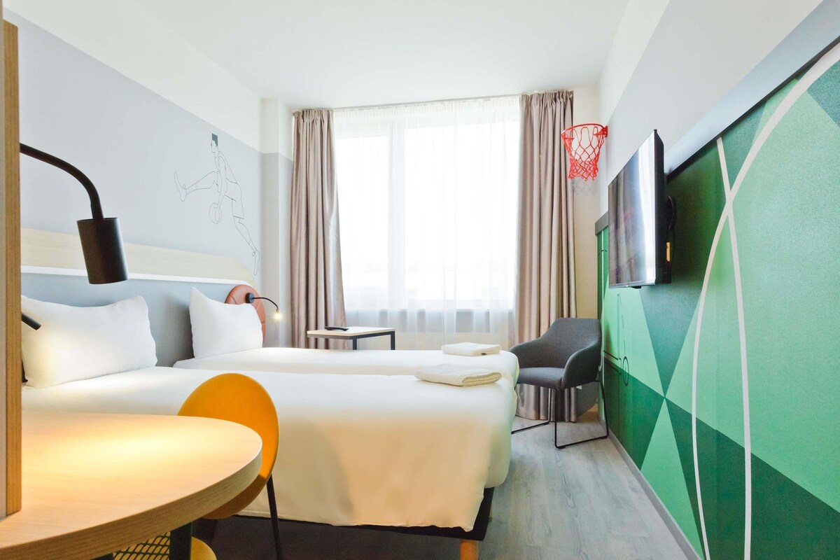 A hotel room with a bed, a desk, a chair, a TV, and a window with curtains.