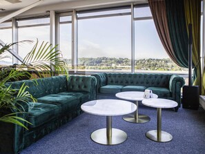 A room with a sectional sofa, two round tables, and large windows with a view. - Ibis Styles Kaunas Centre (Kaunas)