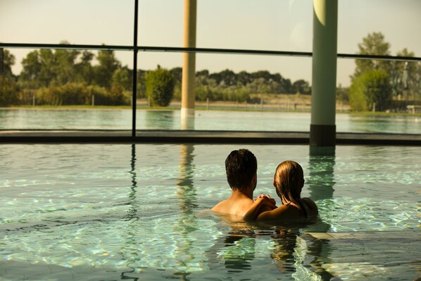 Una pareja en una piscina interior transparente con ventanas amplias y vistas a árboles y un puente.