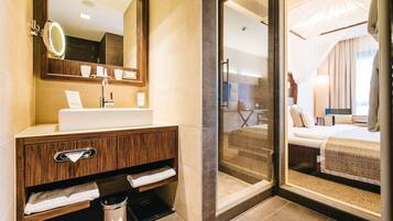 A modern bathroom with a vanity, mirror, and a glass door leading to a bedroom with a bed and curtains.