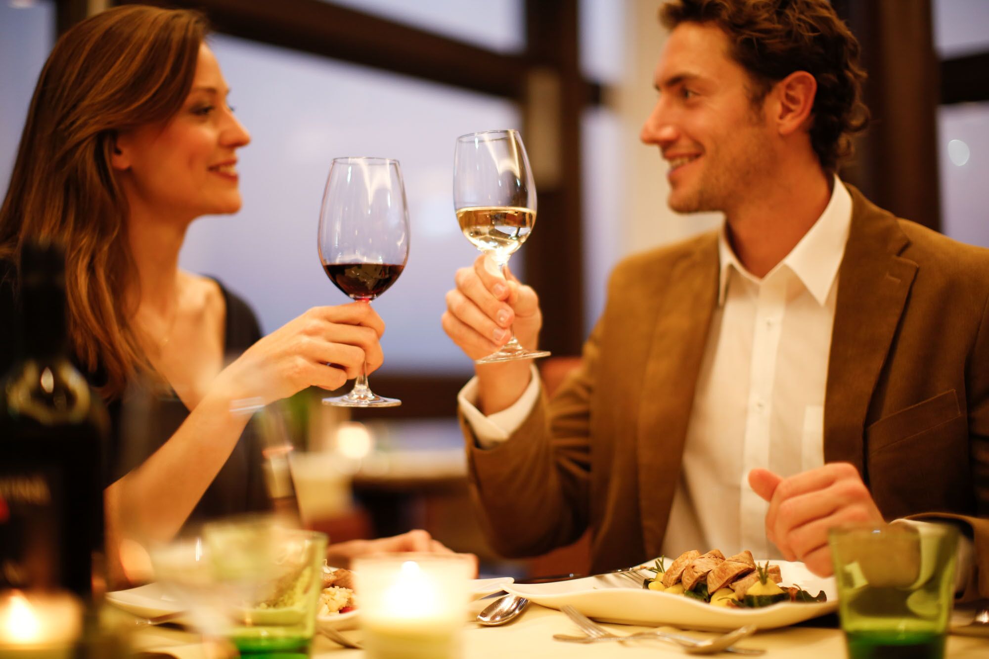 A couple toasting with wine glasses at a dinner table with a candle, surrounded by plates of food and a green glass.