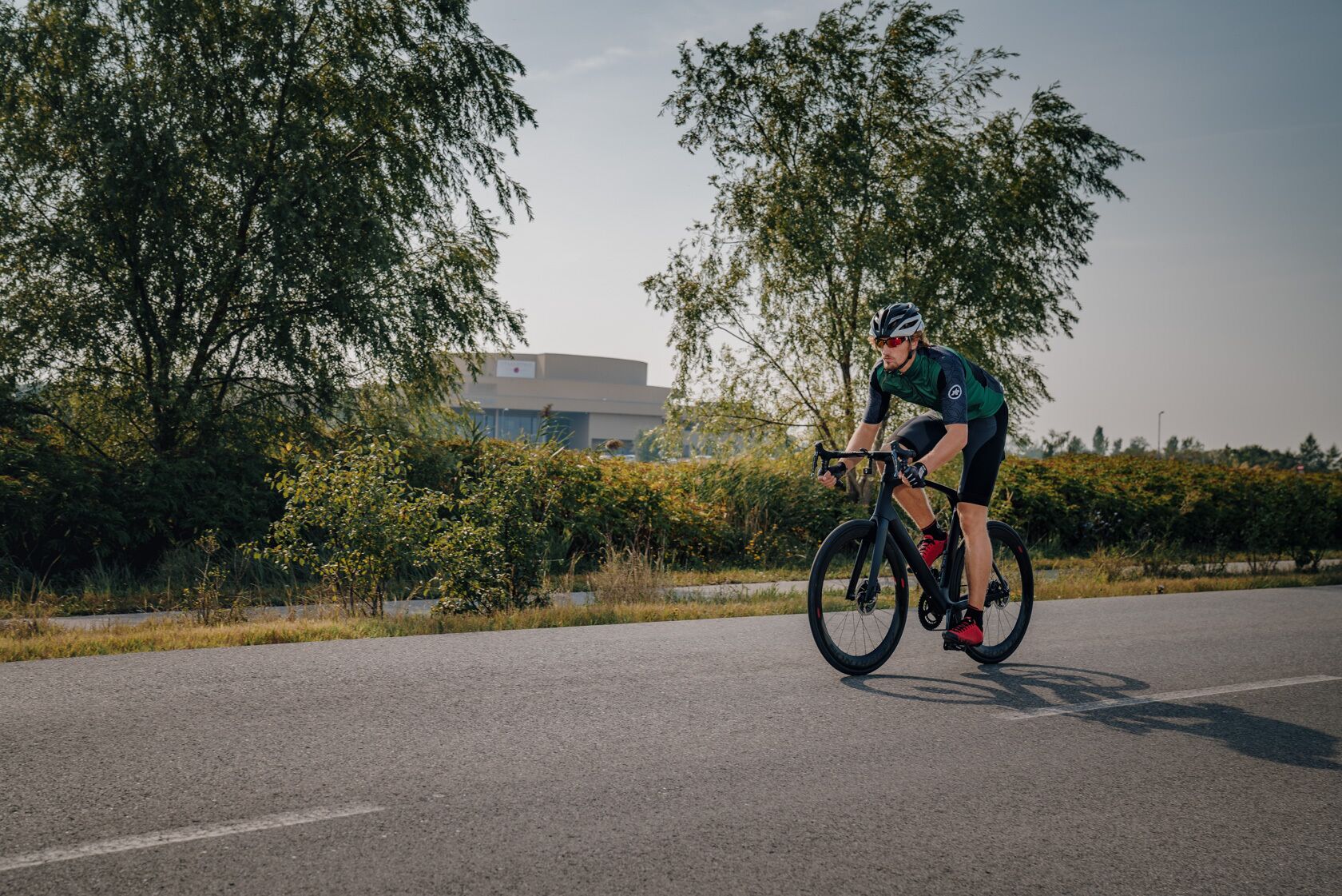 Um ciclista pedala em uma estrada pavimentada, com árvores e um edifício ao fundo.