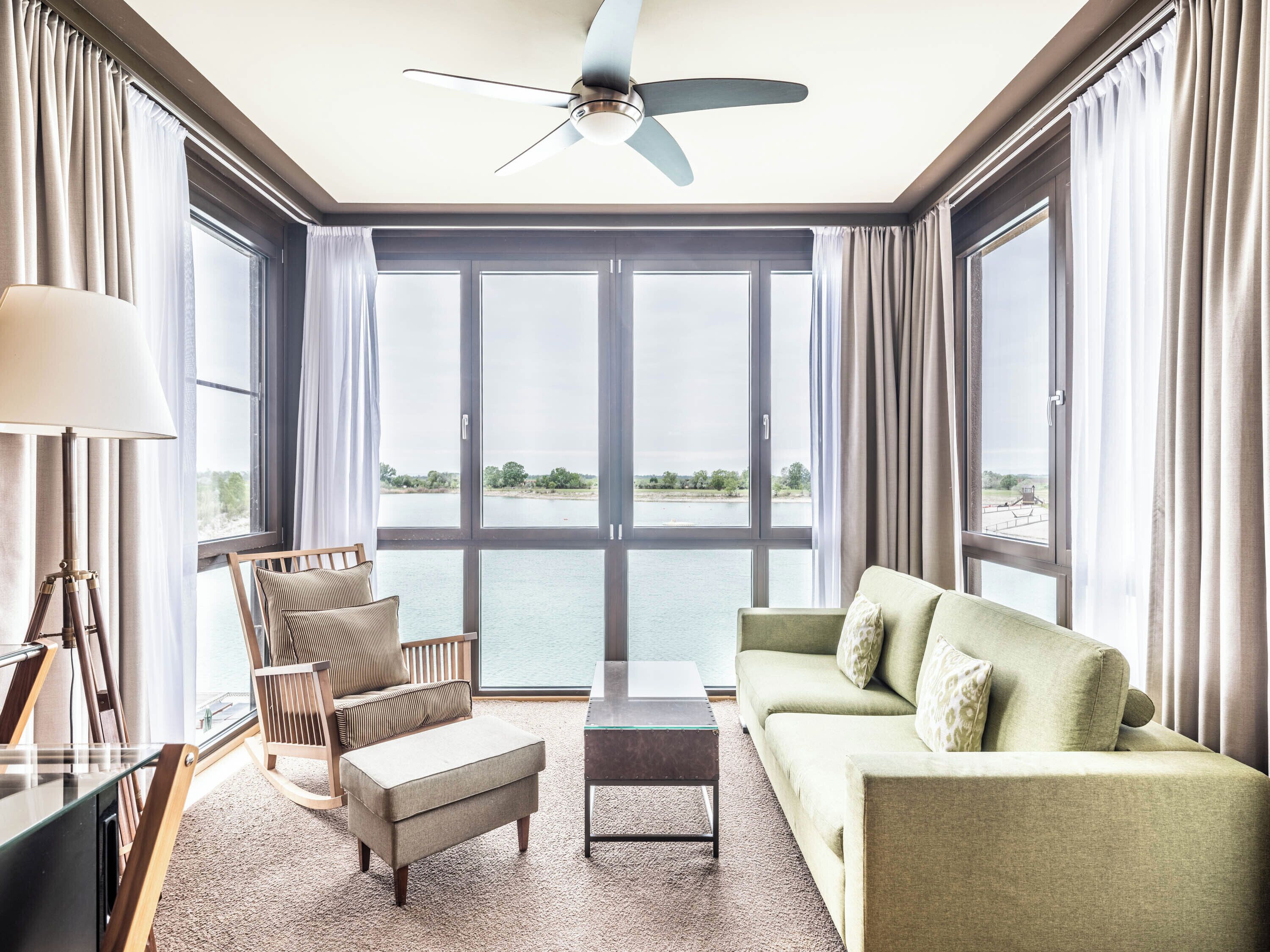 Ein geräumiges Zimmer mit großem Fenster, Deckenventilator, Sofa, Sessel und kleinem Tisch.