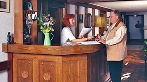 Un mostrador de recepción con encimera de madera, un jarrón con flores y una maleta antigua en el suelo.