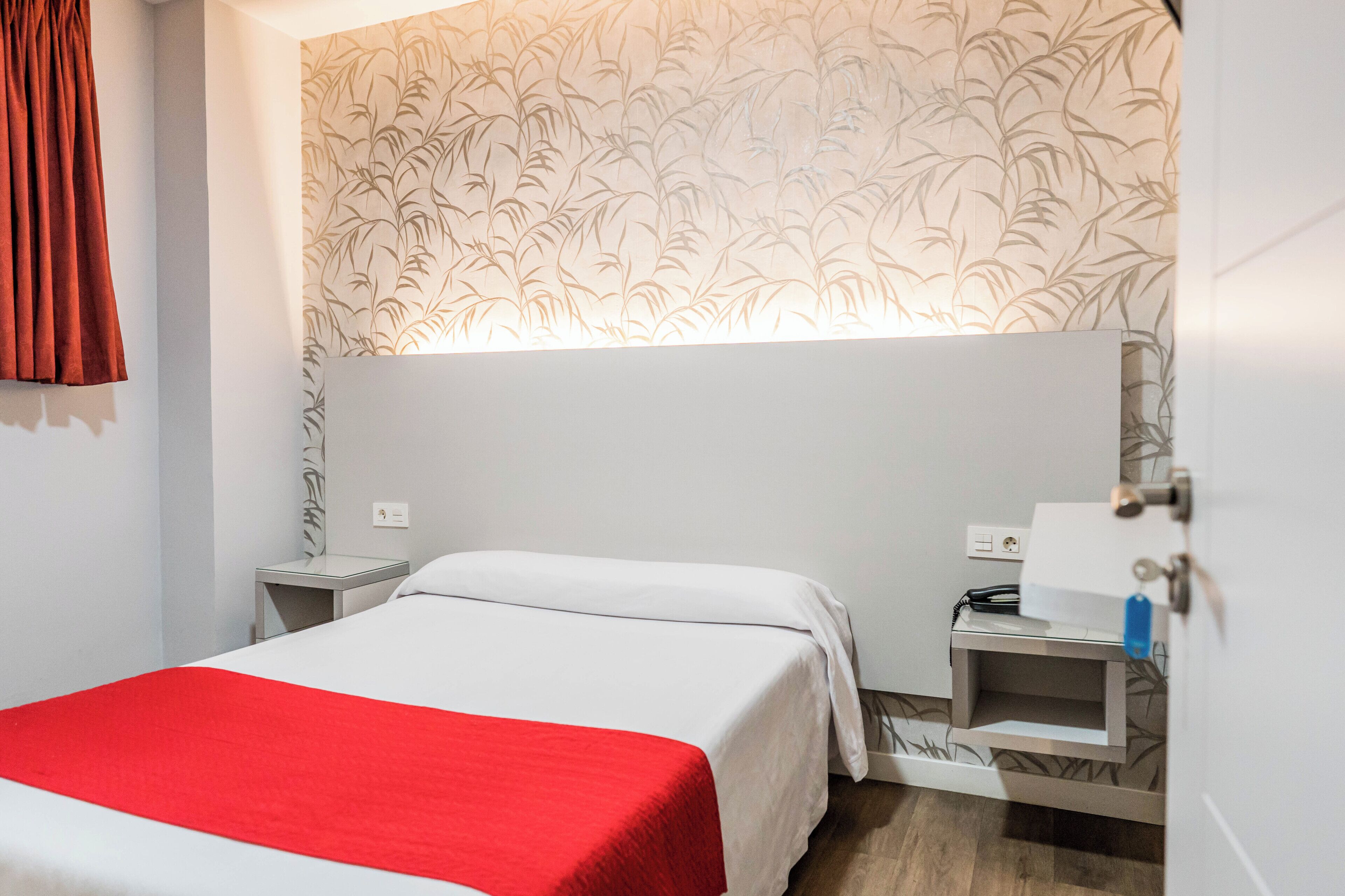 A hotel room with a bed, a nightstand, a desk with a lamp, and a wall with a bamboo pattern.