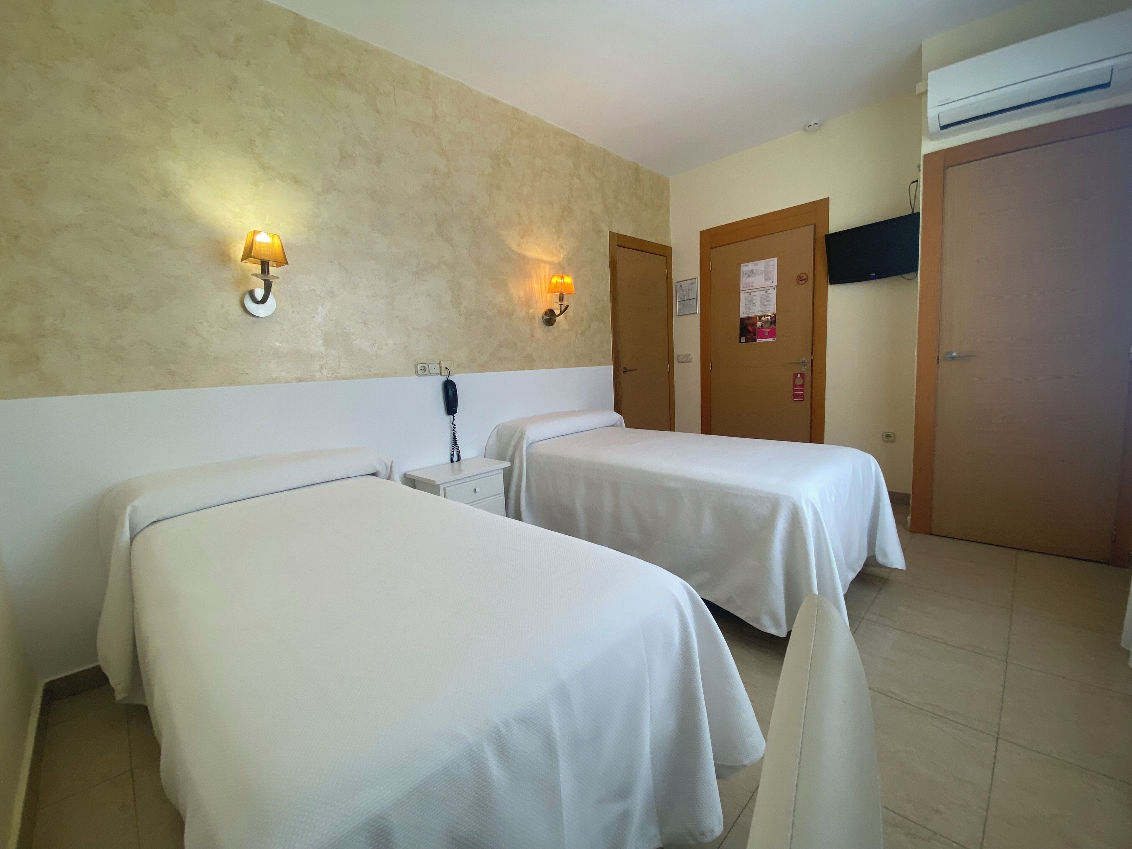 Una habitación de hotel con dos camas individuales, una mesita de noche con lámpara, un teléfono en la pared y un equipo de aire acondicionado.