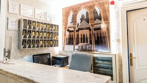 Een receptiebalie met een marmeren blad, een computer en een stoel. Er hangen gecertificeerde lijstjes aan de muur en een groot schilderij in een lijst van een gebouw met bogen.