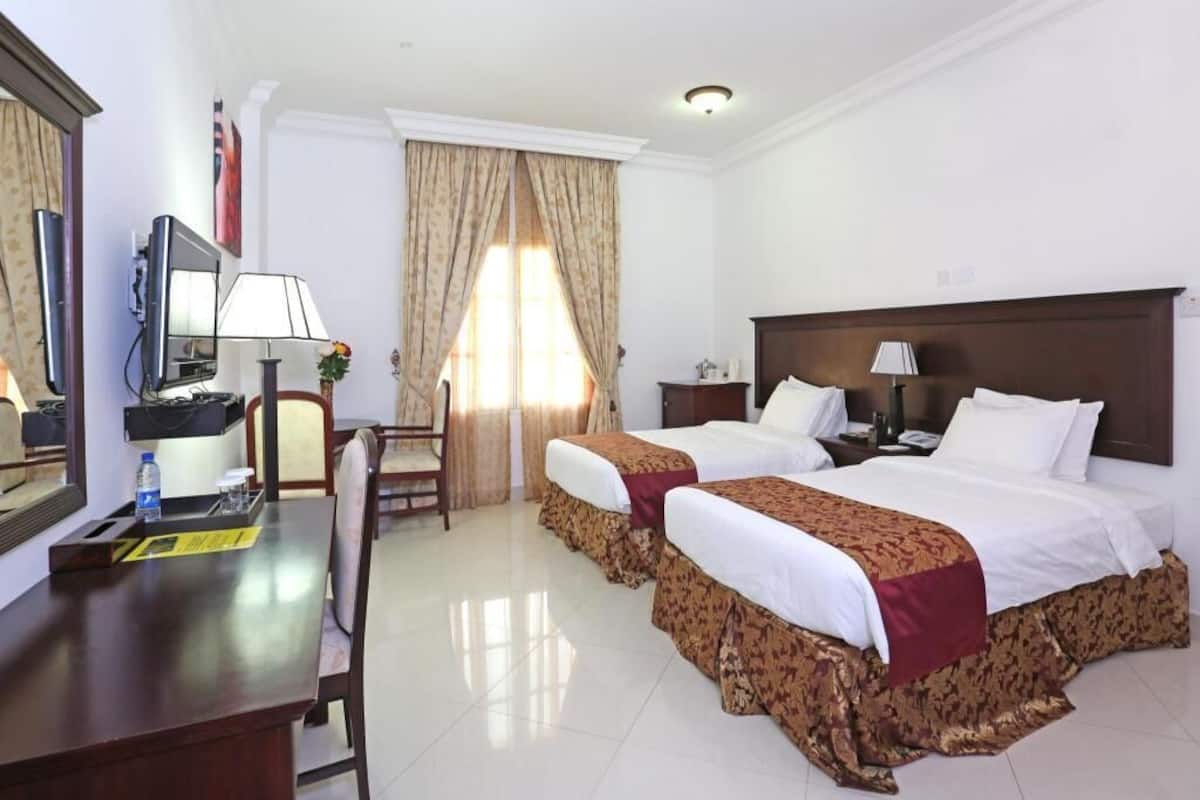 A hotel room with two beds, a desk with a chair, a TV, and a window with curtains.