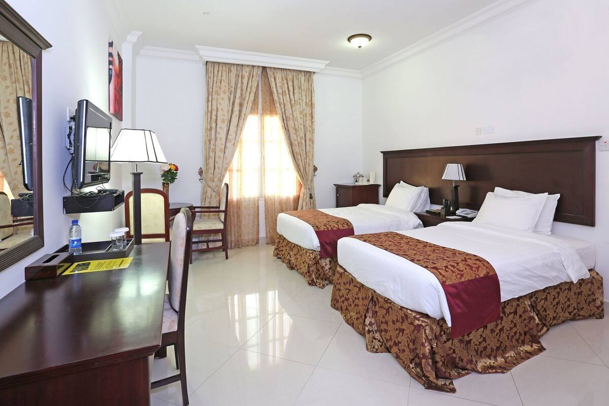A hotel room with two beds, a desk with a chair, a TV, and a lamp.