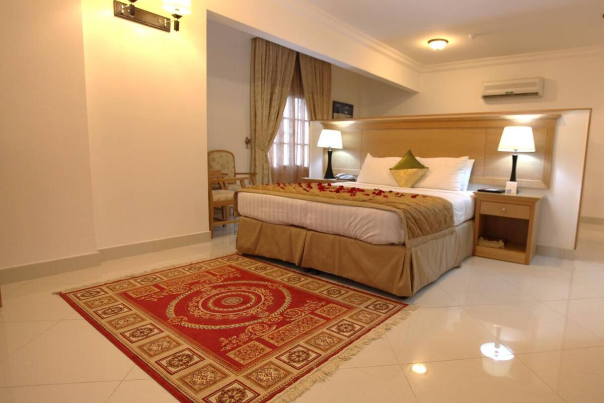 A hotel room with a bed, bedside tables, a lamp, a rug, and an air conditioning unit.