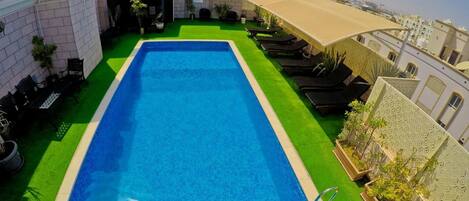 A rectangular swimming pool with a blue interior, surrounded by green artificial turf. There are lounge chairs and a covered seating area with a table and chairs. The pool is adjacent to a building with a beige facade and a decorative wall with intricate patterns.