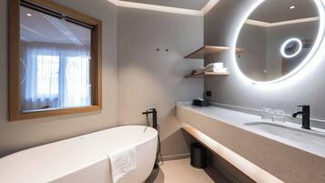 Une salle de bain moderne avec une baignoire sur pieds, un grand miroir avec un éclairage circulaire et une fenêtre ornée de rideaux légers.