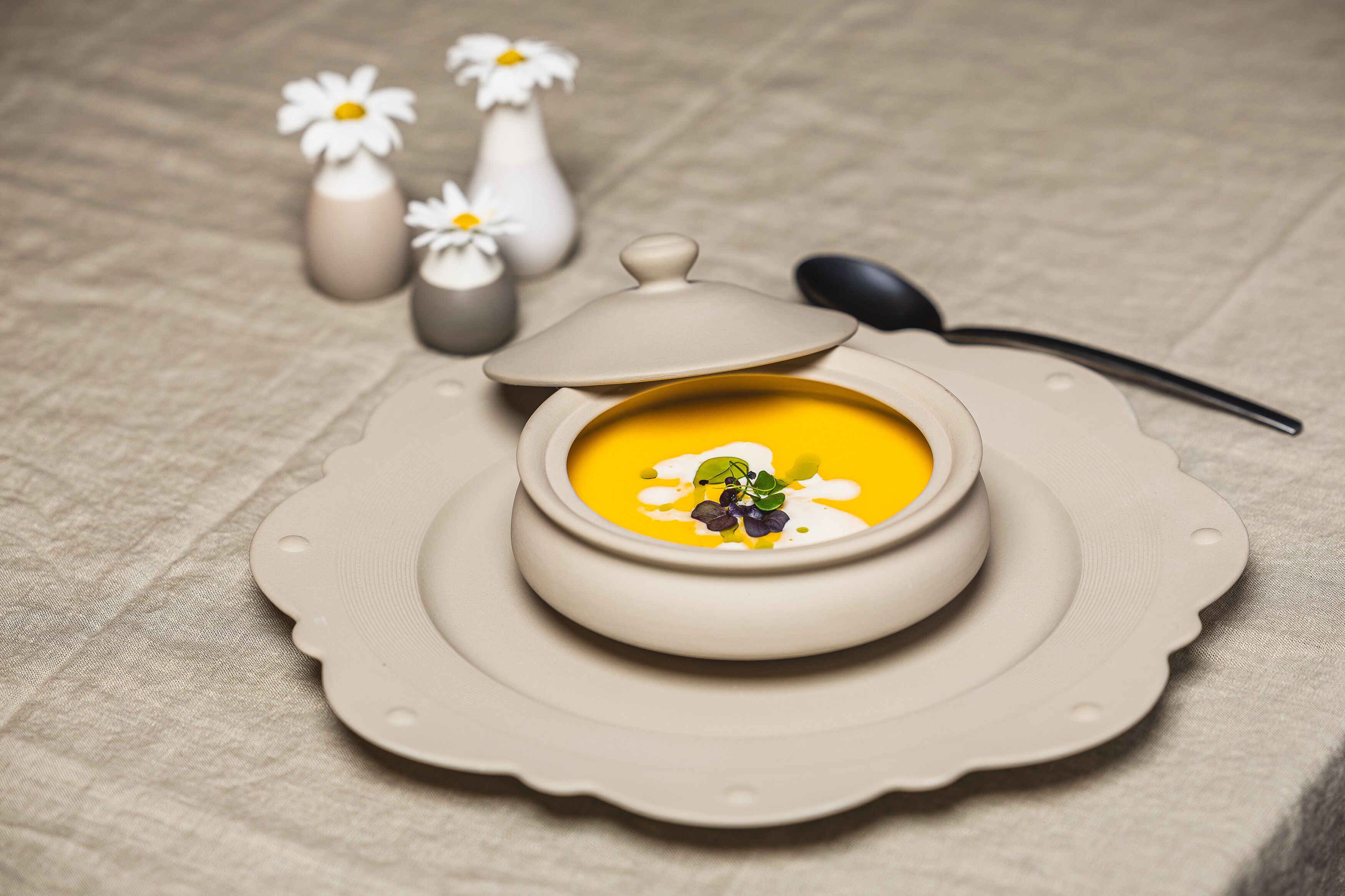 A bowl of soup on a beige plate with a scalloped edge, accompanied by a spoon and two small vases with flowers.
