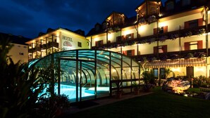 A hotel with a glass-enclosed swimming pool, multiple balconies with flower boxes, and a well-lit exterior at night.
