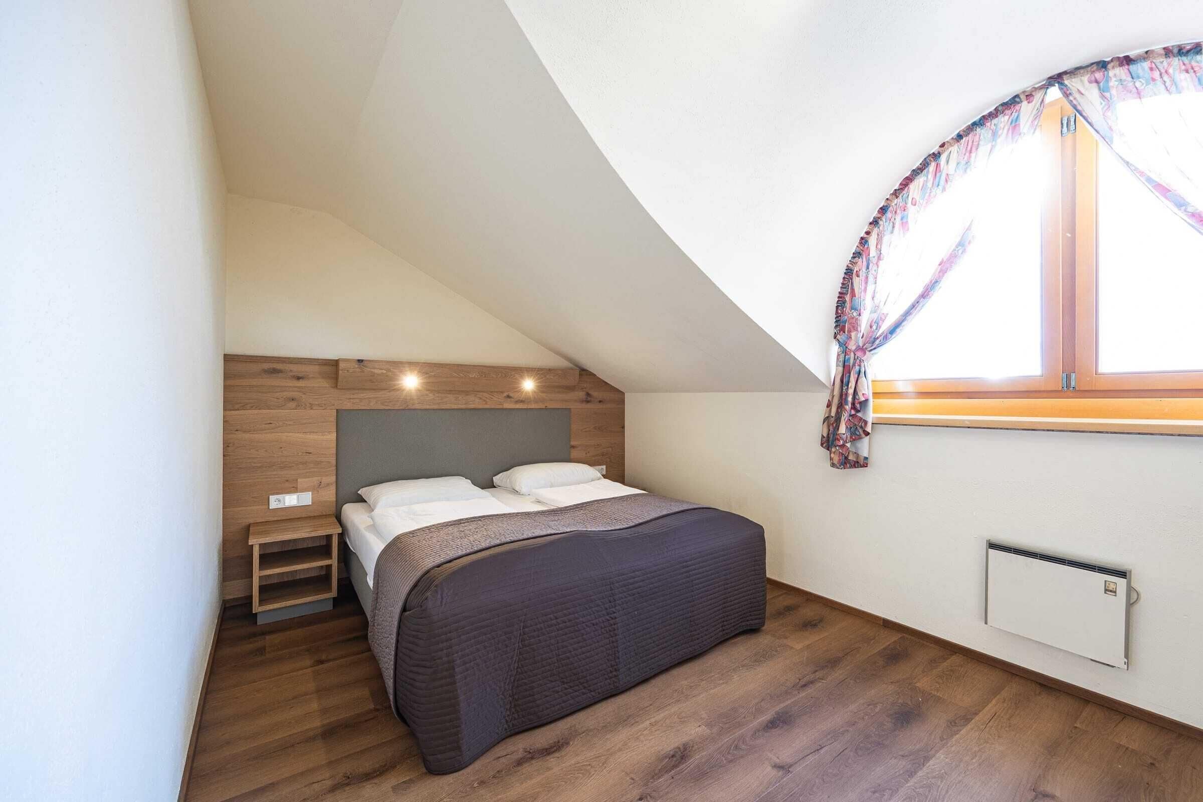 A bedroom with a sloped ceiling, a bed with a dark headboard, a wooden nightstand, a radiator, and a window with a curtain.