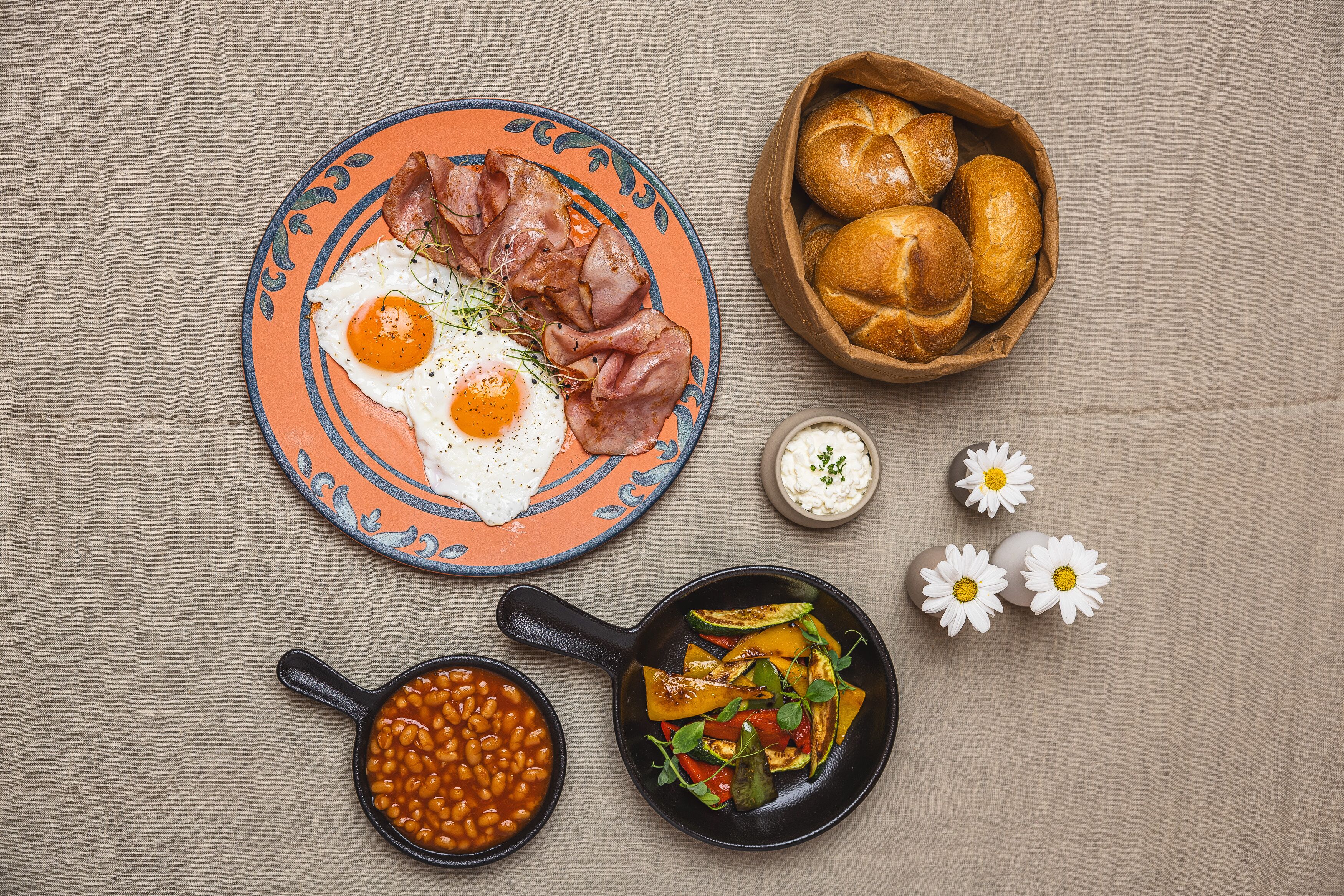 A plate with eggs, bacon, and beans, a skillet with vegetables, and a bowl of rolls on a tablecloth.