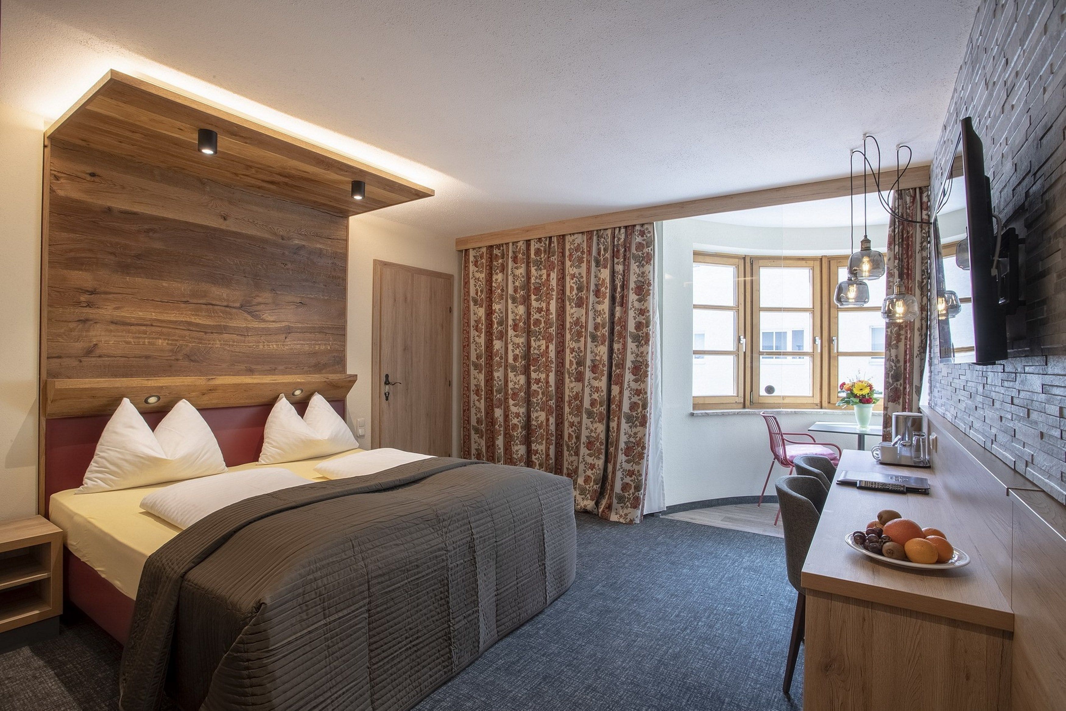 A modern hotel room with a large bed, a wooden headboard, a desk with a fruit bowl, and a TV mounted on the wall.