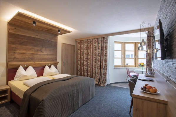 Una habitación de hotel moderna con una cama grande, cabecera de madera, un escritorio con frutero y un televisor montado en la pared.