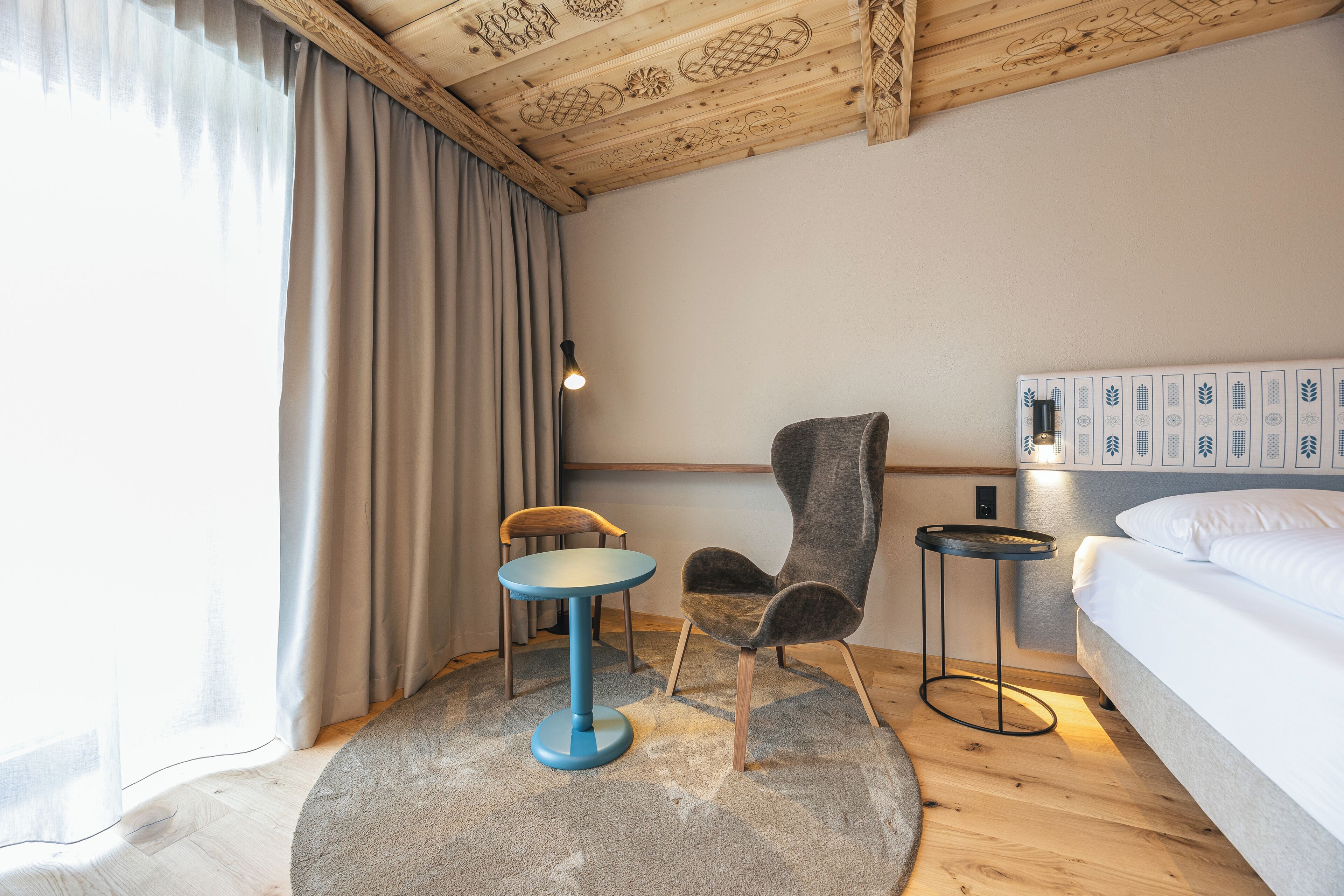 A hotel room with a wooden ceiling, a bed, a round table, a chair, a nightstand, and a wall with a decorative panel.