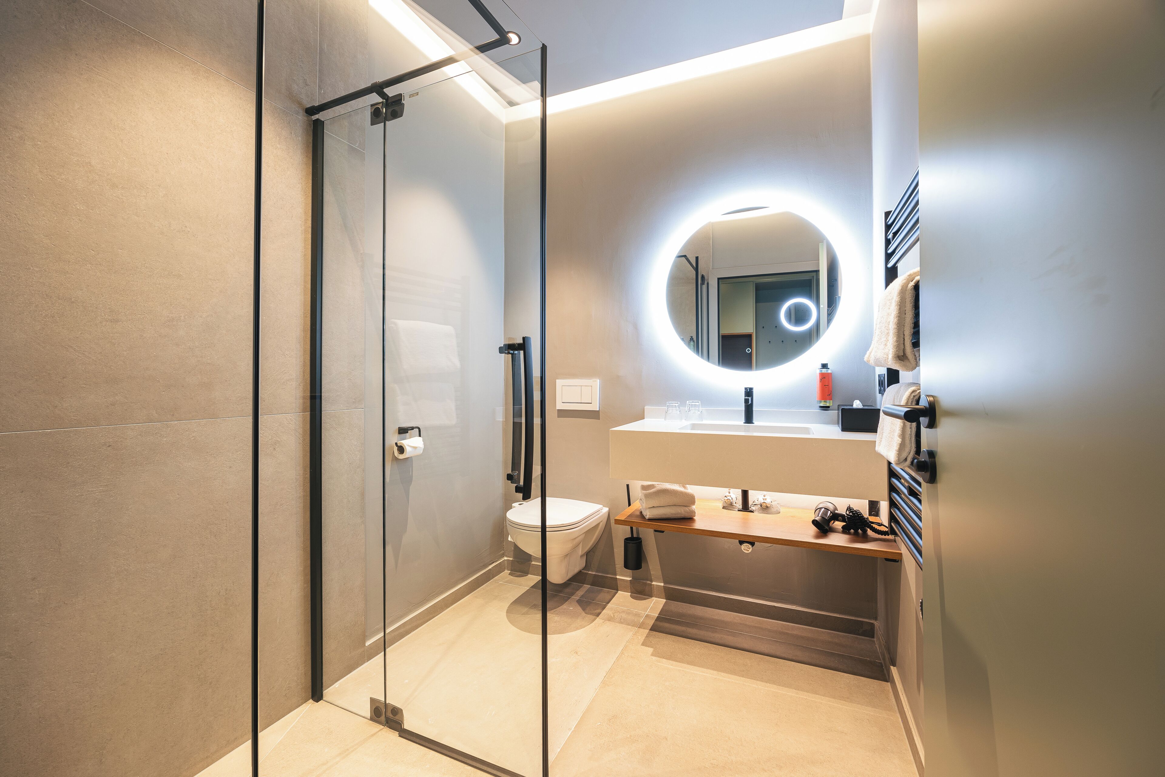 Ein modernes Badezimmer mit einer Duschkabine aus Glas, einem runden Spiegel mit integrierter Beleuchtung, einer Toilette und einem Waschtisch mit Spüle und Handtuchhalter.