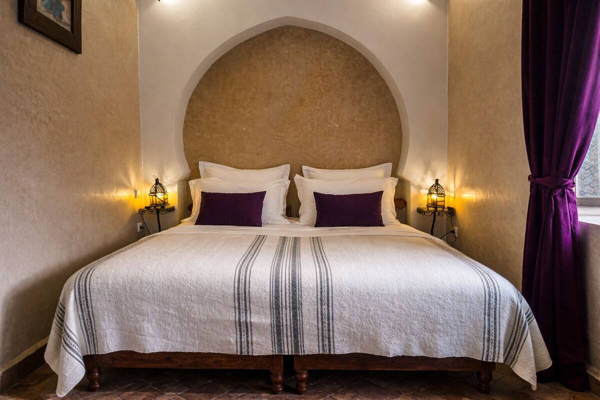 A bed with a white and gray striped bedspread, two white pillows, and two purple pillows. There are two wall-mounted lamps on either side of the bed.