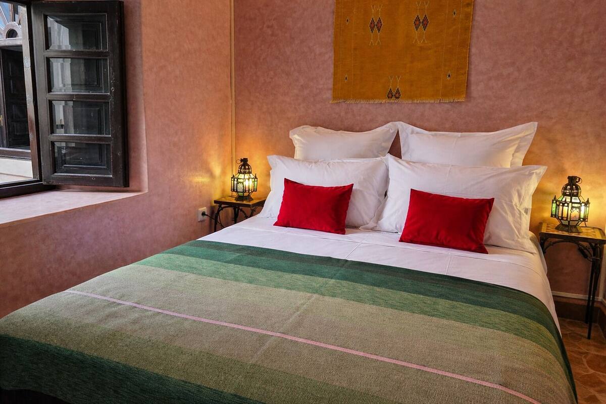 A neatly made bed with white and red pillows, a green striped blanket, and two wall-mounted lanterns on either side.