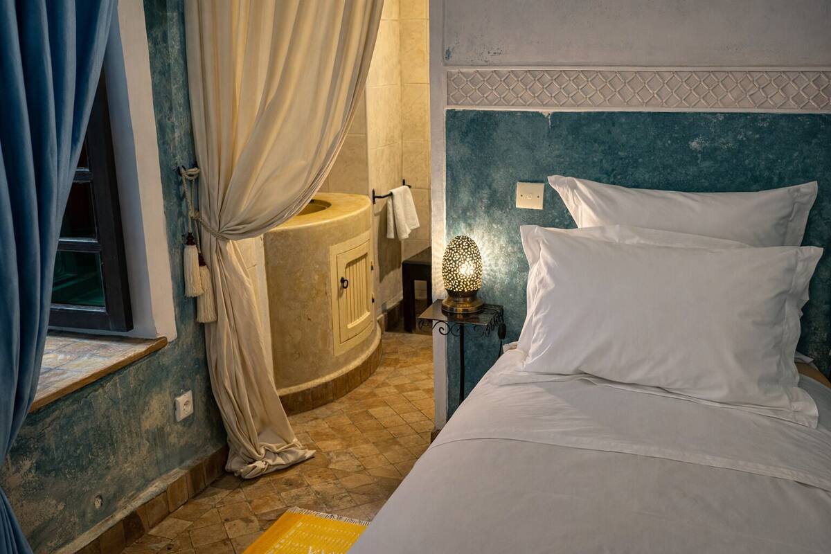 A bedroom with a bed, a nightstand with a lamp, a window with curtains, and a bathroom with a sink and towel rack.