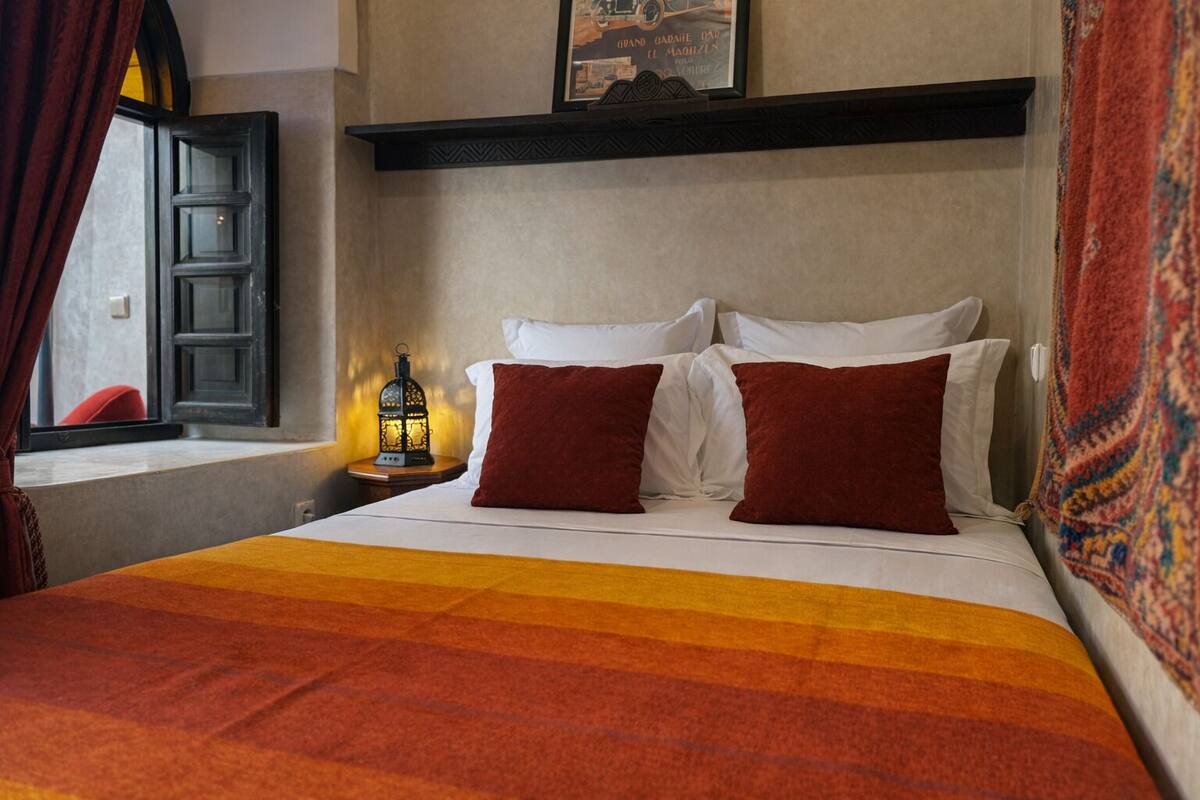 A hotel room with a bed, two red pillows, a nightstand with a lantern, a window with curtains, and a wall-mounted shelf with a framed picture.