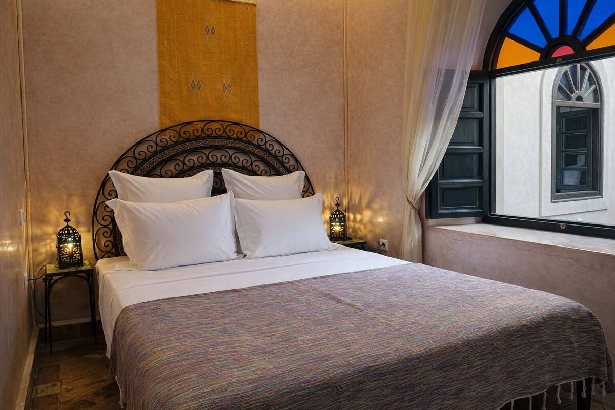 A neatly made bed with white linens and a patterned throw, a decorative headboard, two bedside tables with lantern-style lamps, and a window with a view of a building.
