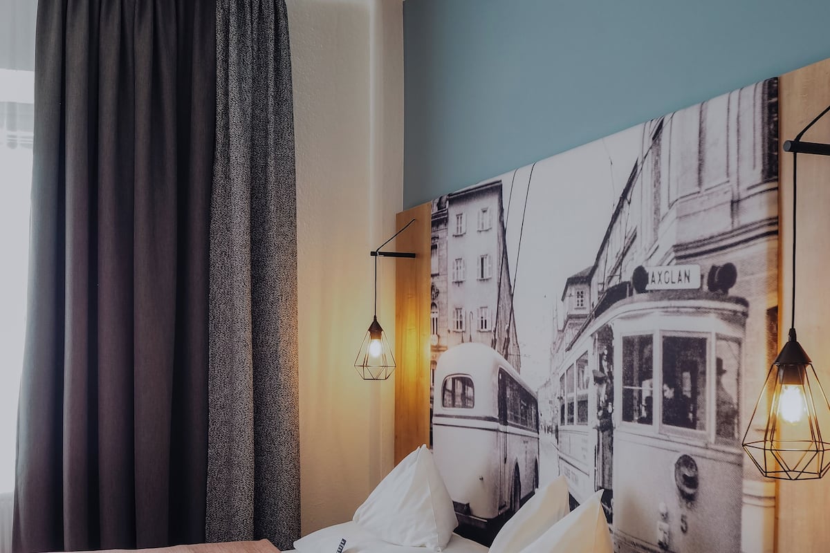 Une chambre d’hôtel avec un lit, une table de chevet, une lampe, un grand tableau représentant un paysage urbain et une fenêtre avec des rideaux.