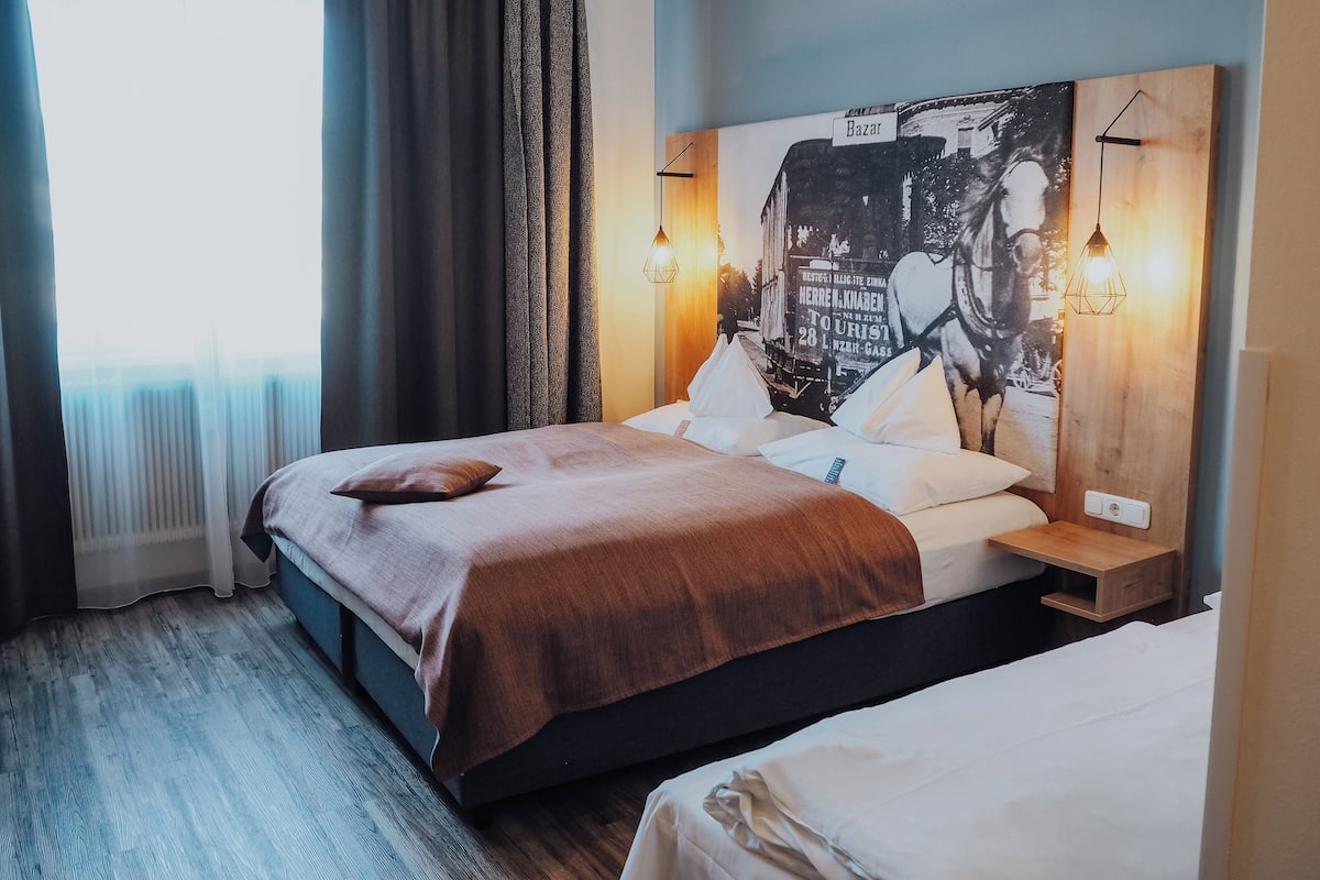 Une chambre d’hôtel avec un lit, une table de chevet, une lampe fixée au mur et une fenêtre avec des rideaux.