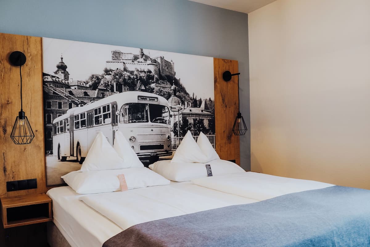 Un lit avec une literie blanche, une couverture bleue et une étagère en bois fixée au mur, sur laquelle se trouve une lampe noire.
