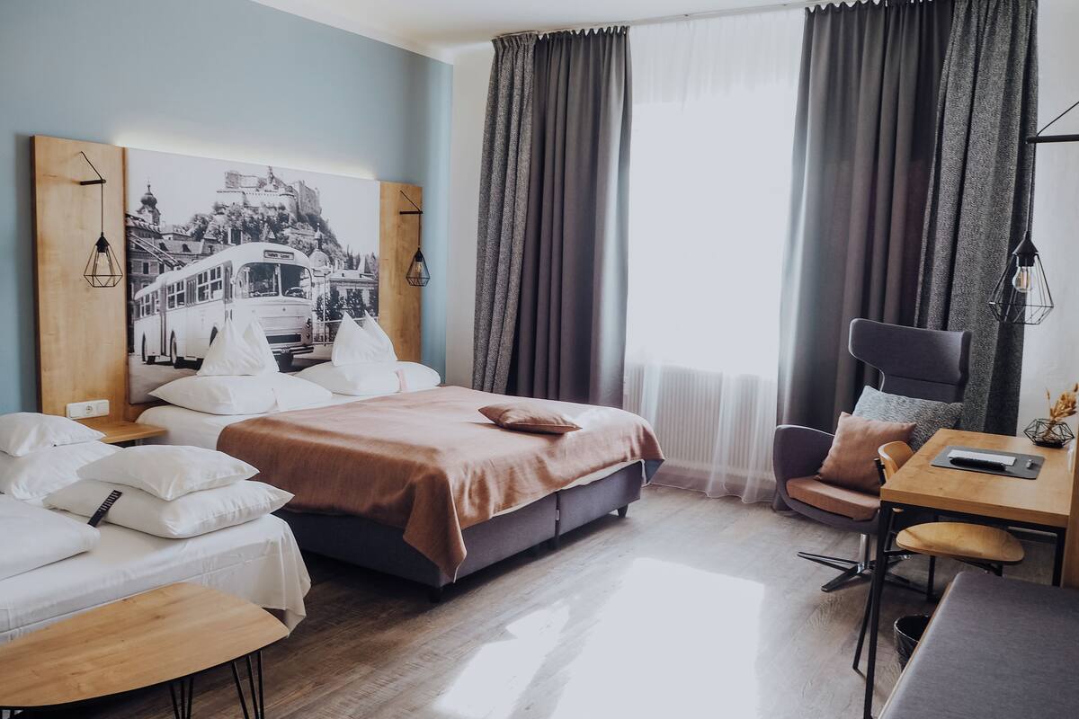 Une chambre d’hôtel comprenant un lit, deux tables de chevet, un bureau avec une chaise et une grande fenêtre avec des rideaux.