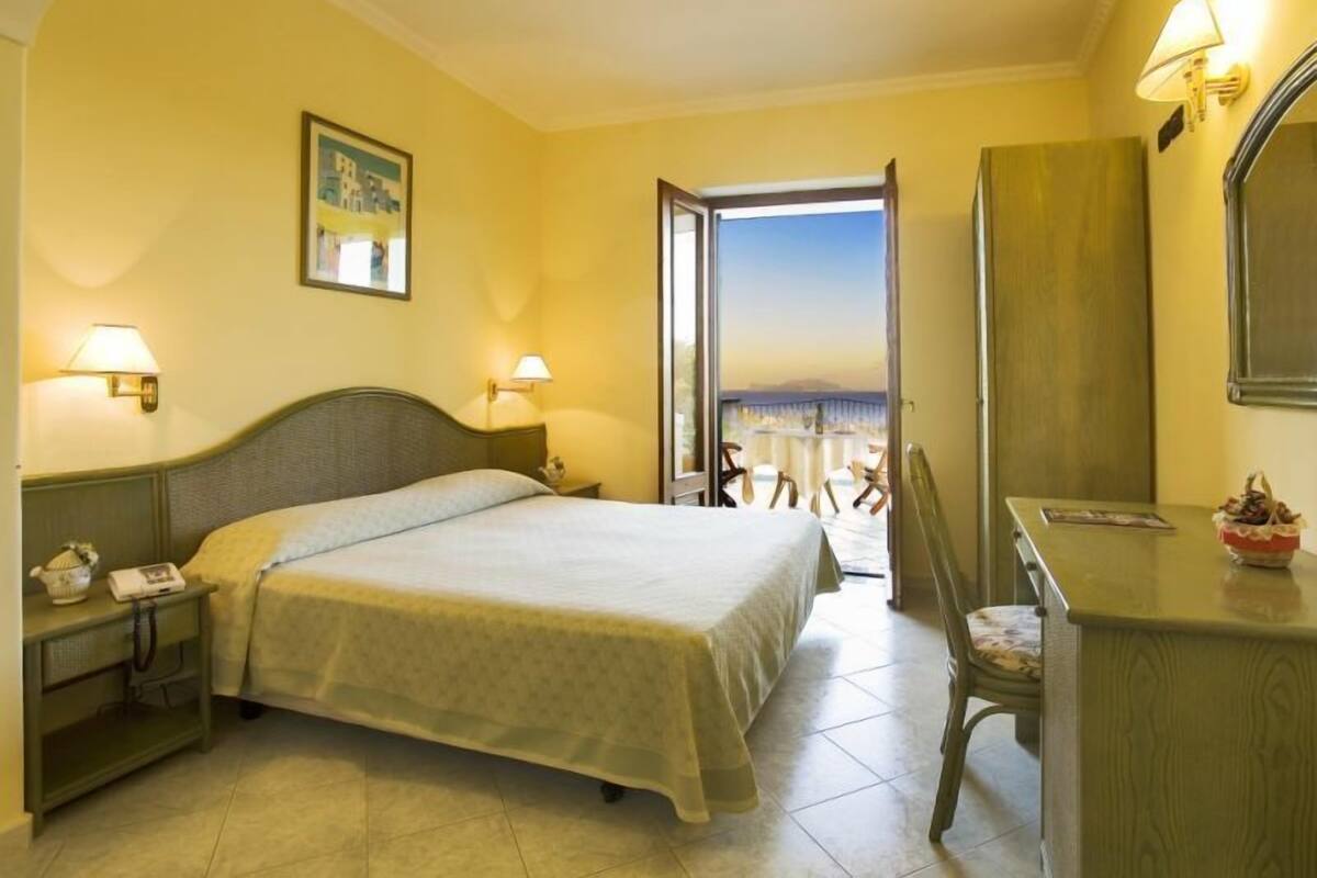 Une chambre d’hôtel avec un lit, des tables de chevet, un bureau et une chaise. Il y a une fenêtre donnant sur la ville.