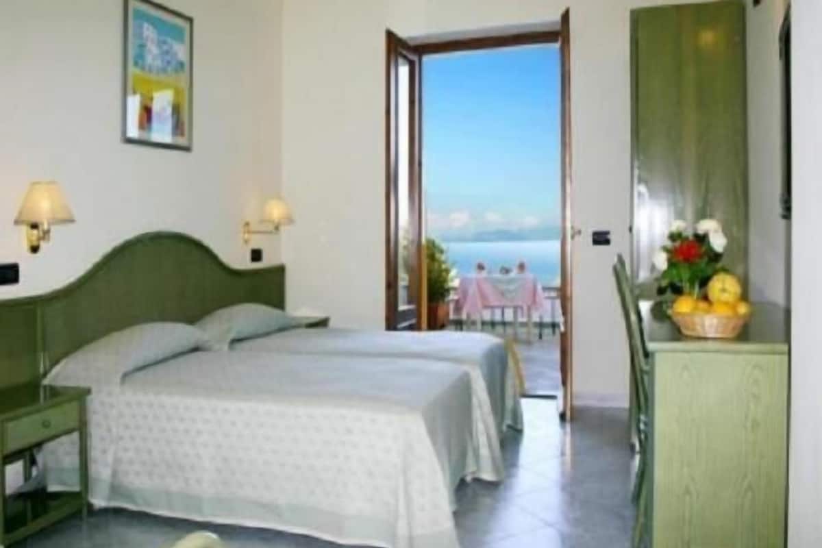 Une chambre d’hôtel avec deux lits, un bureau avec une lampe, une vue sur la mer et un balcon avec une table et des chaises.